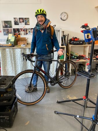 Male cyclist wearing a yellow helmet and holding his bike in the Meadowbank tool library. A stand is visible in the foreground to the right, with shelving and work surfaces in the background.