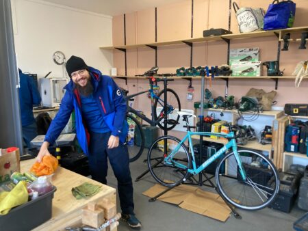 Male cyclist in blue waterproof gear placing something on a worktop surface. In the background is a bicycle with a light blue frame leaning against some shelves, and another black frame bike in a stand.